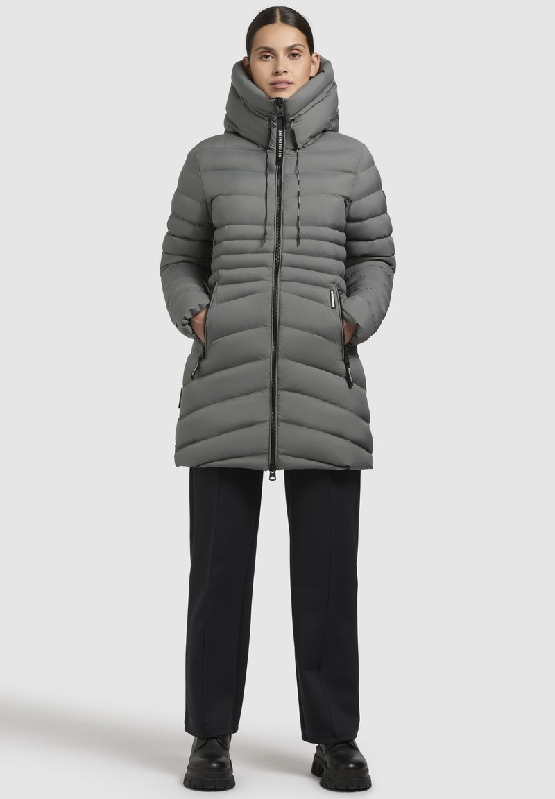 Cappotto imbottito grigio con colletto alto, baffles verticali, chiusura con zip frontale e tasche laterali. Abbinato a pantaloni neri e stivali neri chunky.