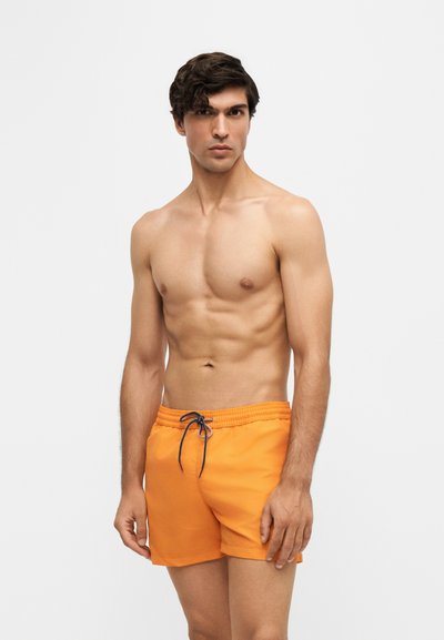 Jeune homme aux cheveux foncés portant un short de bain orange vif avec un cordon noir, debout devant un fond clair uni.