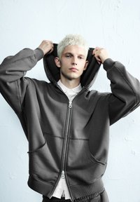 Mladý človek s krátkymi blond vlasmi, oblečený v tmavej mikine na zips cez bielu košeľu, upravuje kapucňu proti pozadiu bílej textúrovanej steny.