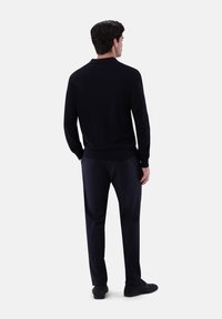 Marineblauer Pullover mit geripptem Saum und Bündchen, hergestellt aus weichem Textil, kombiniert mit dunkel gekleideten Hosen und schwarzen Schuhen, von hinten gezeigt.