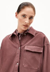 Frau mit zurückgebundenem Haar, die eine mauvefarbene Jacke mit Knöpfen und einer großen Brusttasche über einem gestreiften Hemd mit Kragen trägt.