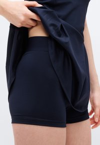 Main soulevant une chemise bleu marine pour révéler un short bleu marine taille haute et serré, porté sur les hanches et le haut des cuisses, sur un fond uni.