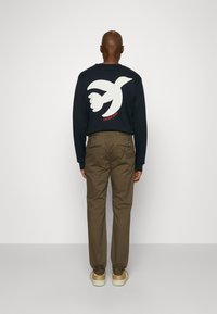 Námornícky crewneck sveter s bielou grafikou vtáka na chrbte, v kombinácii s khaki nohavicami a béžovými topánkami. Hladká tkanina, voľný strih.
