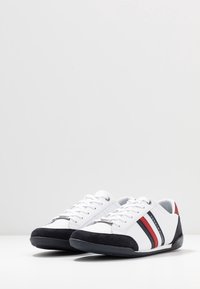 Ett par vita sneakers med marinblå och röda ränder, svarta tåskydd, vita snören och "Tommy Hilfiger"-logotyp på sidan.