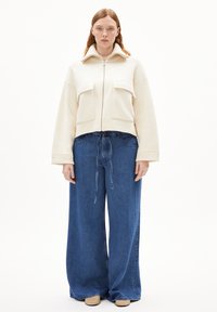 Cream zip-up jacket with two front pockets, paired with loose, high-waisted blue denim jeans and beige slip-on shoes.