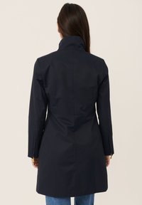 Marineblå trench coat med høj krave, skræddersyet pasform og vertikal søm-detaljering. Har lynlåse ved manchetterne og et glat, tekstureret stof.