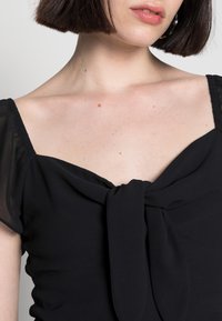 Svart tygblus med fyrkantig halsringning, puffärmar och en vriden rosett detalj på framsidan. Slät textur och figurnära design.