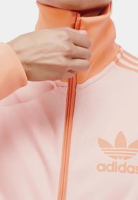 Svijetlo ružičasta atletska jakna s visokim ovratnikom i zatvaranjem na zip; sadrži narančasti zip i usklađeni detalj na ovratniku. Adidas logo na prsima.