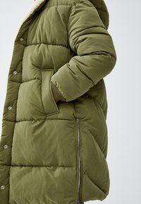 Chaqueta acolchada de color verde oliva con cierre de cremallera, secciones grandes acolchadas, bolsillos laterales y una capucha suave forrada de felpa.