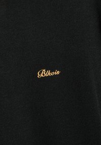 Μαύρος πλεκτός ιστός με μικρή καλλιγραφική κεντημένη ένδειξη "Blkvis" σε κίτρινο νήμα κεντραρισμένη στην επιφάνεια.