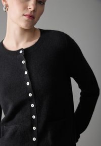 Black cardigan made of soft material, featuring a crew neck, white button closure, and two front pockets, with a fitted silhouette.