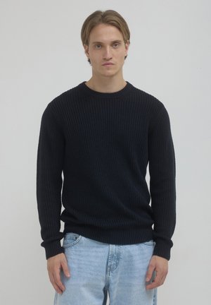 Junger Mann mit hellbraunem Haar, der einen schwarzen Strickpullover und hellblaue Jeans trägt und vor einem schlichten hellen Hintergrund steht.