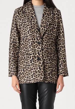 Vrouw die een blazer met luipaardprint draagt met twee zwarte knopen, over een grijs geribd topje en zwarte leren broek, staand tegen een effen achtergrond.