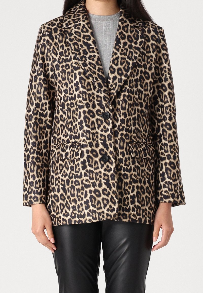 Femme portant un blazer à imprimé léopard avec deux boutons noirs, par-dessus un haut gris côtelé et un pantalon en cuir noir, debout devant un fond uni.