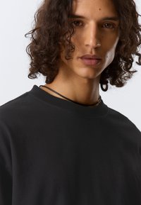 Tricou negru din bumbac, cu guler rotund, mâneci scurte și o textură netedă. Modelul are părul creț și poartă o colier negru.