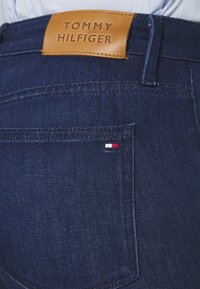 Tummansiniset farkut, joissa on ruskea nahkatagi, jossa lukee "TOMMY HILFIGER". Pienen punaisen, valkoisen ja sinisen lipputunnuksen embloma takataskussa.