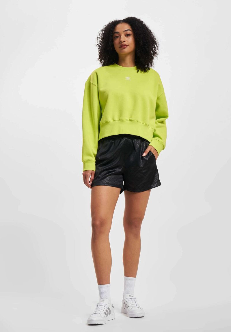 Sweatshirt court vert lime avec un petit logo blanc, porté avec un short noir texturé, des chaussettes blanches et des baskets blanches.