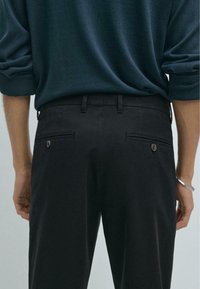 Schwarze Hosen mit geradem Schnitt, ausgestattet mit zwei Gesäßtaschen und Knopfdetails, kombiniert mit einem locker sitzenden dunkelgrünen Pullover.