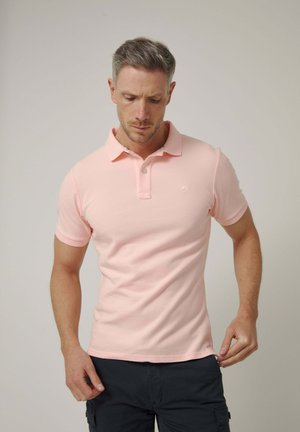 Hombre con pelo corto y gris, que lleva una camiseta polo rosa clara y pantalones oscuros, mirando hacia abajo mientras ajusta el dobladillo de la camisa.