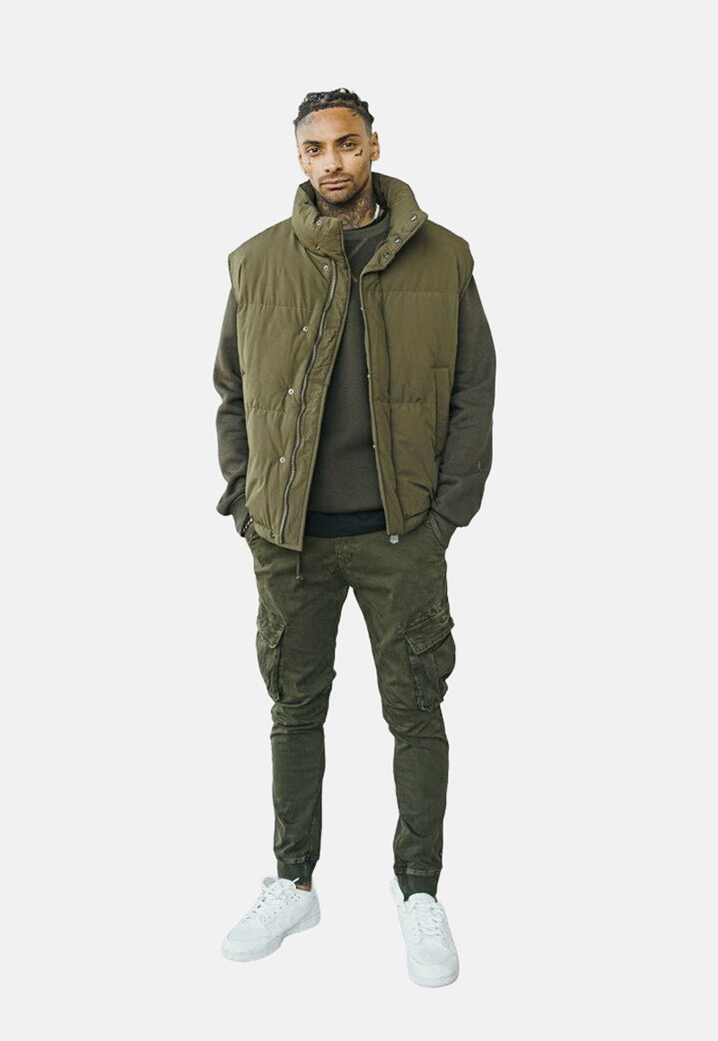 Gilet matelassé vert olive sur un pull sombre, associé à un pantalon cargo et des baskets blanches. Les caractéristiques incluent des fermetures à boutons et des poches.