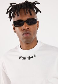 Hvid langærmet skjorte med "New York" trykt i sort gotisk skrift på brystet. Sorte rektangulære solbriller og flettet frisure synlig.