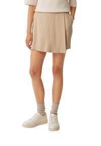 Beige skort med en blød, glat tekstur; den har et plisseret design og en halvcirkelform, parret med hvide sneakers og beige ankelstrømper.