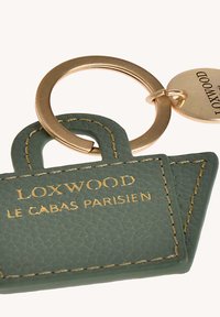 Porte-clés en cuir vert en forme de sac avec un texte en or embossé « LOXWOOD » et « LE CABAS PARISIEN », attaché à un anneau en métal doré.