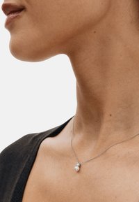 Hõbedast kettkaelakee, millel on ümar pärl ja väike vääriskivist ripats, kantakse kaela lähedal musta topiga.