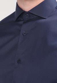Marineblaues Button-up-Hemd mit glatter Textur, klassischem Kragen und zwei schwarzen Knöpfen. Der Stoff wirkt leicht und minimal zerknittert.