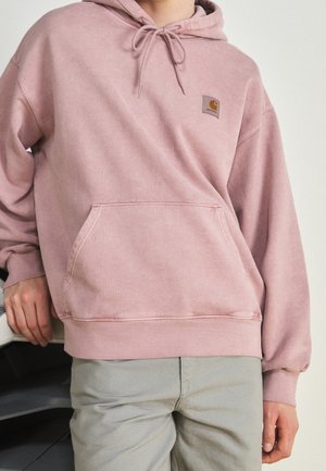 Person iført en støvet lyserød Carhartt hættetrøje med frontlomme og beige bukser, der læner sig op ad en hvid bil foran en neutral baggrund.