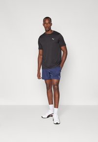 Black short-sleeve athletic shirt with a logo, paired with navy shorts. Model wears white sneakers with red accents and white socks.