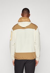 Let jakke i creme og brun, med hætte, elastiske manchetter og et Columbia-logo på ryggen. Textureret stof med farveblokering.