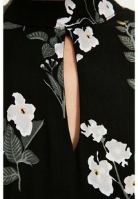 Zwarte stoffen blouse met een keyhole halslijn en een bloemenpatroon in wit en grijs, met groene bladeren en gele accenten.