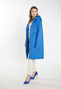 Palton lung captusit, de culoare albastră, cu glugă, fermoar frontal și capse. Purtați deasupra unei bluze albe și a unor pantaloni crem, asortați cu tocuri înalte albastre.