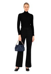 Vrouw in een zwarte turtleneck en zwart-witte geblokte broek, met een donkerblauwe handtas met een gouden kettinghengsel, in zwarte schoenen, staande op een witte achtergrond.