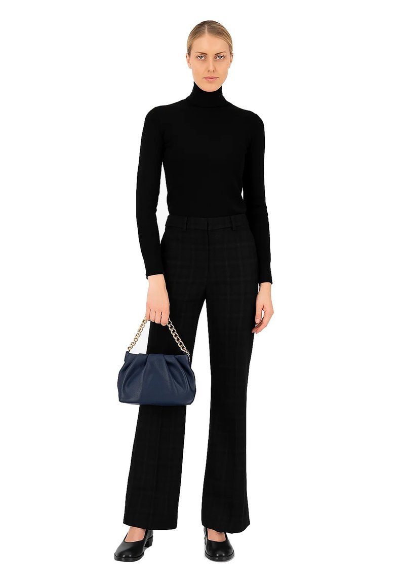 Vrouw in een zwarte turtleneck en zwart-witte geblokte broek, met een donkerblauwe handtas met een gouden kettinghengsel, in zwarte schoenen, staande op een witte achtergrond.