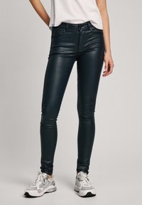 Úzké džíny v tmavé teal barvě, vyrobené z lesklého materiálu. Mají klasický design s pěti kapsami, zipový pas a úzký střih. Jsou nošeny s bílými teniskami.