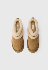 Zapatillas de ante marrón con un cuello de piel sintética crema, forma redondeada y patrones circulares. Diseñadas para la comodidad con una suela gruesa.