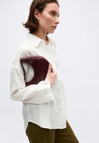 Νέα γυναίκα με λευκό πουκάμισο και ελαιώδες παντελόνι κρατάει μια μικρή σκούρο κόκκινη τσάντα με υφή κάτω από το μπράτσο της, στραμμένη προς τα αριστερά μπροστά σε απλό φόντο.