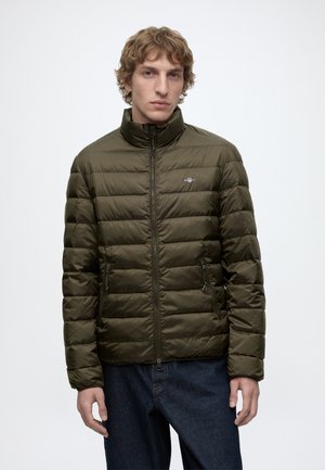 Junger Mann mit lockigem Haar, der eine olivgrüne gesteppte Jacke mit Reißverschluss und dunkelblaue Jeans trägt, vor einem schlichten weißen Hintergrund steht.