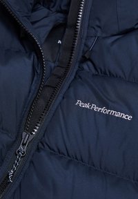 Marineblå pufferjakke med lynlås, syede sektioner og "Peak Performance" branding i sølvbroderi på forsiden.
