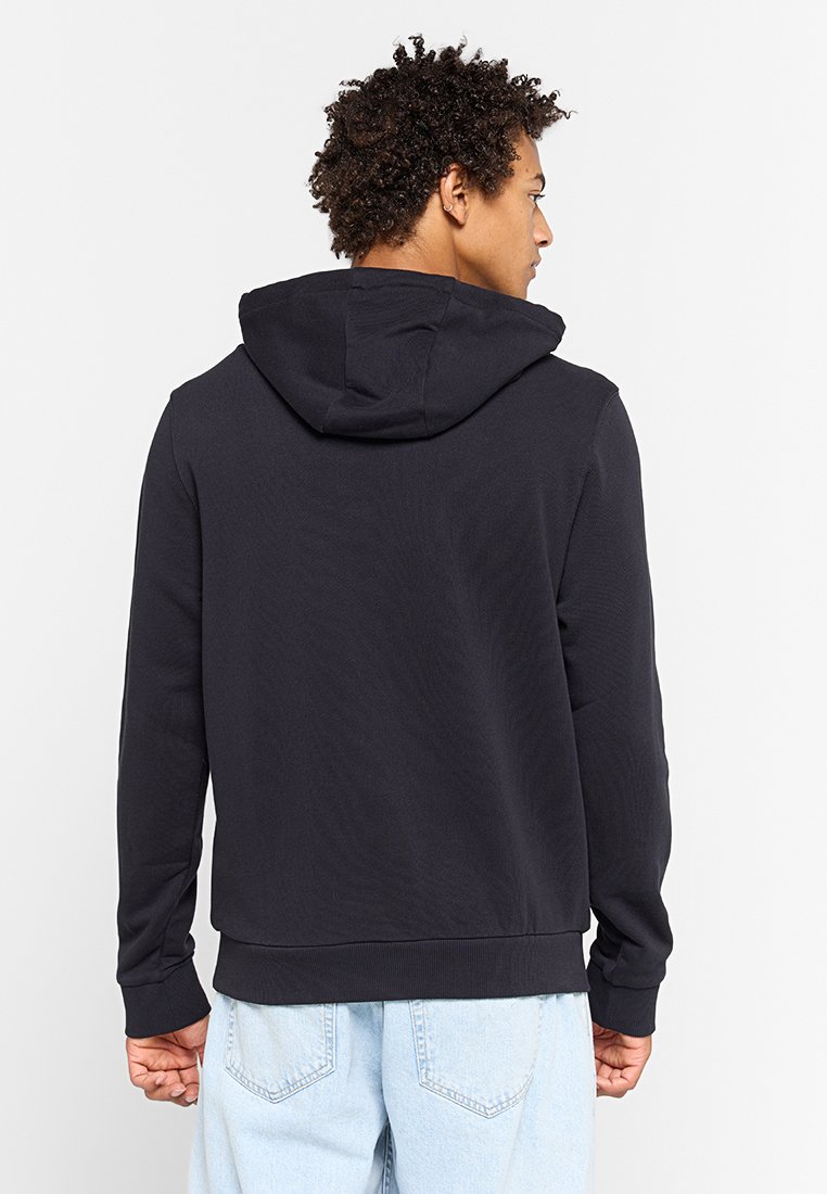 Granatowy hoodie z kapturem na sznurku, z prążkowanymi mankietami i dołem, charakteryzujący się gładką, miękką fakturą. Noszony z jasnoniebieskimi dżinsami.