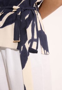 Granatowy i kremowy pasek w wzory związany w talii na białych spodniach, ukazujący fakturę materiału i detal bocznego rozcięcia.