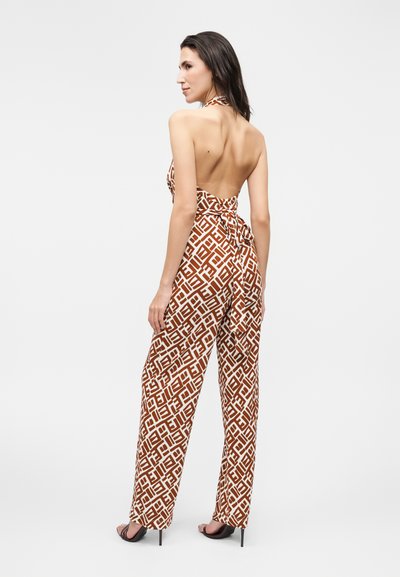 Γυναίκα που φοράει αμάνικη, ανοιχτή στην πλάτη, καφέ και λευκή ολόσωμη φόρμα με φαρδιά πόδια, στέκεται πλαγίως με ψηλοτάκουνα σανδάλια σε μονόχρωμο φόντο.