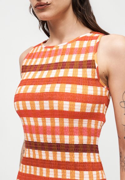Γυναίκα που φοράει αμάνικο εφαρμοστό φόρεμα με οριζόντιο και κάθετο καρό μοτίβο σε πορτοκαλί, κόκκινες, κίτρινες, ροζ και λευκές αποχρώσεις.