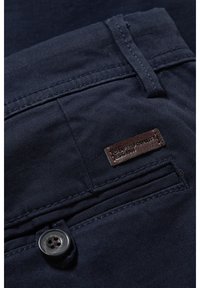 Poche de pantalon bleu foncé avec bouton noir et petite étiquette en cuir marron cousue au-dessus de l’ouverture de la poche.