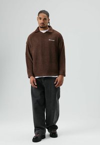 Polaire marron à col avec un logo brodé, associée à un jean noir ample et des chaussures noires à lacets.