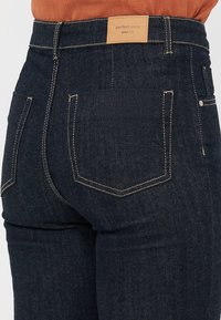 Mørkeblå denim jeans med høy midje, med kontrasterende gul søm og et skinnmerke på baklivet. To baklommer.