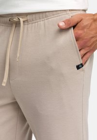 Close-up of beige pants with drawstring waist, side pocket, and hand resting inside pocket against white background.