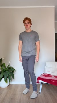 Ung man med röd hår som bär en grå t-shirt, mörka byxor och beige slip-on skor som står inomhus nära en växt och en vit stol med en röd filt.